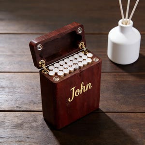 Puede incluir: Un estuche de cigarrillos de madera marrón oscuro con el nombre "John" en letras doradas. El estuche está abierto, revelando cigarrillos. Un difusor de cerámica blanca con cañas está en el fondo.