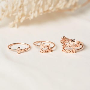 May include: Three gold rings with names engraved on them. The rings are stacked on top of each other. The names on the rings are Maya, Scarlett, and Sarah. The ring on the bottom has the name Sarah on it. The ring in the middle has the name Scarlett on it. The ring on the top has the name Maya on it.