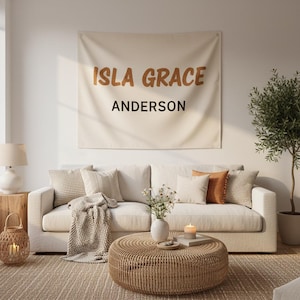 Puede incluir: Un salón con un sofá crema, cojines y una mesa de centro tejida. Un tapiz de pared muestra "ISLA GRACE ANDERSON" en letras marrones. Un pequeño árbol y una lámpara completan la escena.