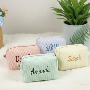 Könnte beinhalten: Vier Kosmetiktaschen aus Cord in Pastellfarben: Rosa, Blau, Grün und Gelb. Jede Tasche hat einen Reißverschluss und einen gestickten Namen. Die Taschen sind rechteckig mit einem Streifenmuster.