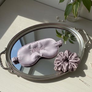 May include: A lavender satin sleep mask embroidered with the name "Olivia" and a matching scrunchie rest on a silver mirrored tray. The tray has ornate handles and is set on a white surface, with a vase of greenery in the background.