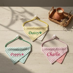 May include: Three personalised dog bandanas in yellow, mint green, and pink, each embroidered with a name. The bandanas feature striped patterns and are attached to elastic bands with white fasteners. A small candle and basket are in the background.