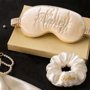 May include: A champagne-colored satin sleep mask with the name Nancy embroidered in gold thread, featuring an elastic strap, paired with a matching white satin scrunchie embroidered with the name Rellen in gold thread, resting on a box.