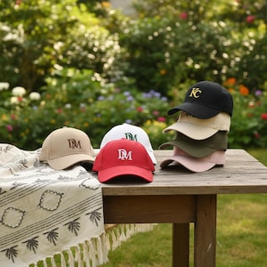 Könnte beinhalten: Eine Auswahl an Baseballkappen in verschiedenen Farben, darunter Schwarz, Beige, Grün und Rosa, ist auf einem Holztisch gestapelt. Weitere Kappen in Beige, Weiß und Rot sind ebenfalls zu sehen. Jede Kappe hat gestickte Buchstaben, einige zeigen "KC" und andere "DM".