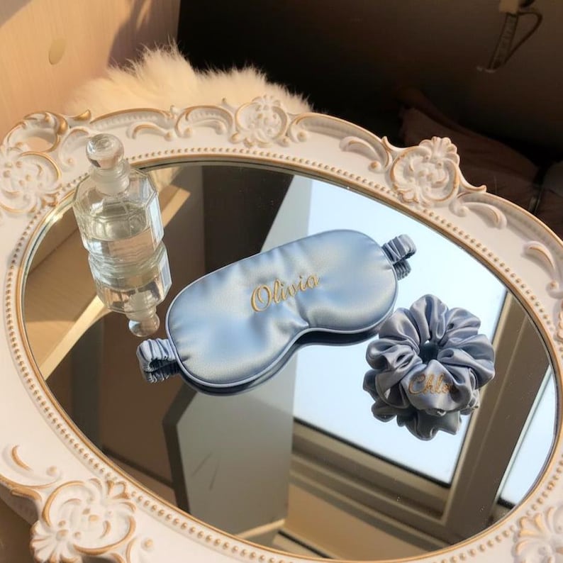 K&ouml;nnte beinhalten: Eine hellblaue Schlafmaske aus Satin und ein passendes Scrunchie mit goldgesticktem Text auf einer verspiegelten Oberfl&auml;che. Die Schlafmaske und das Scrunchie tragen den Namen "Olivia" aufgestickt. Eine klare Glasparf&uuml;mflasche befindet sich ebenfalls auf dem Spiegel.
