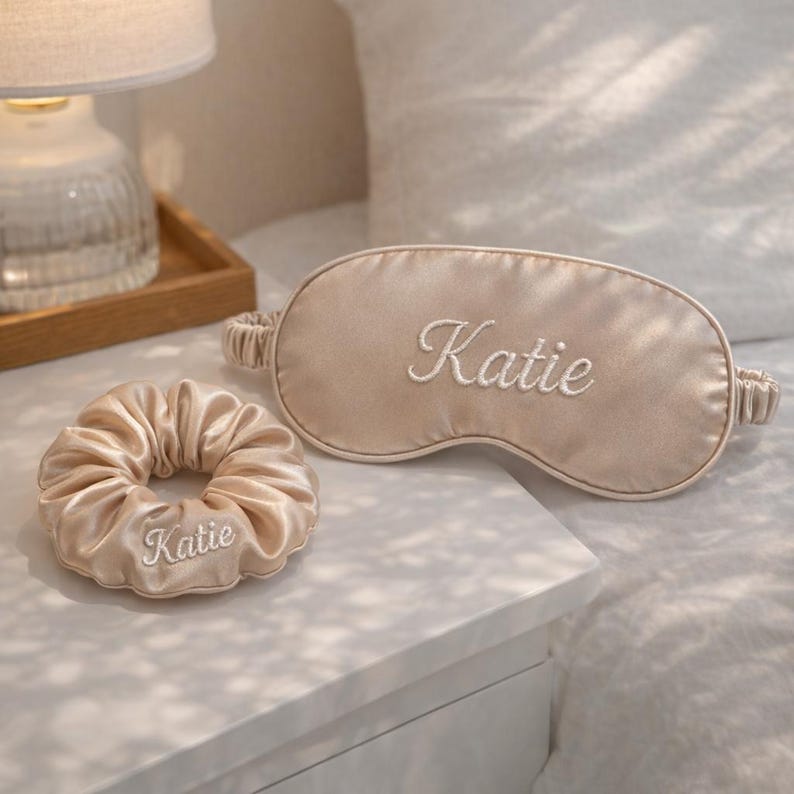 K&ouml;nnte beinhalten: Eine beige Schlafmaske aus Satin und ein passendes Scrunchie, beide mit dem Namen "Katie" in wei&szlig;er Schrift bestickt. Die Schlafmaske hat eine ovale Form mit einem Gummiband. Das Scrunchie ist rund und gerafft.