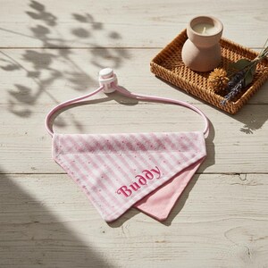 May include: A pink and white striped dog bandana embroidered with the name "Buddy" in pink script. The bandana is attached to a pink adjustable collar. A candle and dried flowers are in the background.