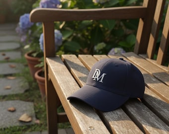 May include: A navy blue baseball cap with a curved brim and a white embroidered "DM" logo. The cap is resting on a wooden bench in an outdoor setting, with a blurred background of flowers and greenery.