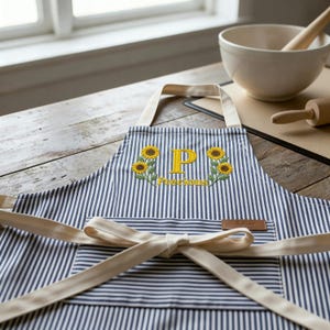 Puede incluir: Delantal a rayas azules y blancas con lazo y correas color crema. El delantal presenta girasoles bordados y la letra "P" con la palabra "Precious" en amarillo. Una pequeña etiqueta de cuero marrón está cosida en el delantal.