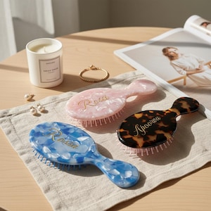 Könnte beinhalten: Drei ovale Haarbürsten in Rosa, Blau und Schildpattmuster. Jede Bürste hat einen Griff und den Namen "Rossi" oder "Apoorva" in goldener Schrift. Die Bürsten liegen auf einem neutralfarbenen Tuch.