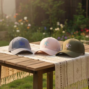 Könnte beinhalten: Drei Baseballkappen in Grau, Rosa und Olivgrün liegen auf einem Holztisch. Jede Kappe hat gestickte Buchstaben: ein "M" auf der grauen Kappe und "KC" auf den rosa und grünen Kappen. Die Kappen werden auf einem dekorativen Tischläufer präsentiert.