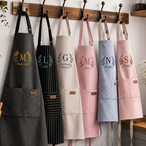 Könnte beinhalten: Sechs gestreifte Schürzen hängen an einer Holzleiste, jede mit einer individuellen Stickerei aus Initial und Name. Die Farben umfassen Anthrazit, Schwarz, Beige, Rosa, Hellblau und Hellrosa, alle mit einer Vordertasche und verstellbaren Riemen.