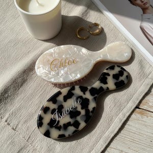 May include: Two personalized hairbrushes. One is white with a pearlescent finish and the name "Chloe" in gold script. The other is black and white with a spotted pattern and the name "Isabelle" in white script. Gold hoop earrings and a candle are also visible.