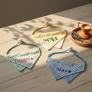 May include: Three dog bandanas with adjustable elastic straps. One is yellow and white striped with "MAX" embroidered in green. Another is mint green and white striped with "Poppy" embroidered in pink. The third is blue and white striped with "Max" and a paw print.