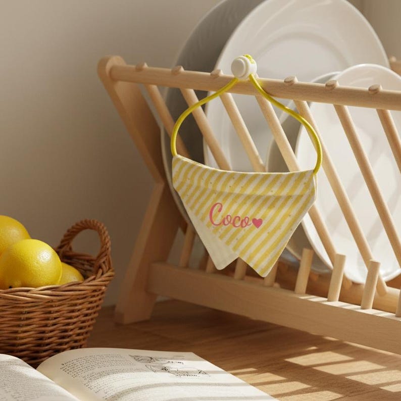 May include: A yellow and white striped bandana with the name "Coco" in pink script and a pink heart. The bandana hangs from a yellow loop attached to a wooden dish rack. A basket of lemons and a book are in the foreground.