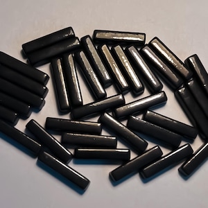 May include: A collection of small, rectangular, black beads scattered across a white surface. The beads have a glossy finish, reflecting light. The image is a close-up, highlighting the texture and shape of the beads, suitable for jewelry making or crafts.
