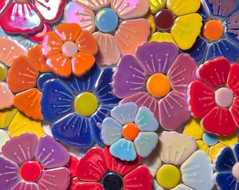 Op de afbeelding: Een verzameling kleurrijke keramische bloemvormige tegels in diverse tinten rood, oranje, geel, blauw, paars en roze. Elke tegel heeft vijf afgeronde bloemblaadjes met reliëflijnen en een contrasterend cirkelvormig midden met een glanzende afwerking.