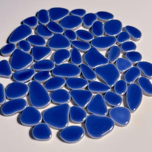 May include: A collection of glossy, cobalt blue ceramic pebbles arranged in a circular pattern. The smooth, rounded shapes vary in size and are set against a white background, creating a visually appealing mosaic.