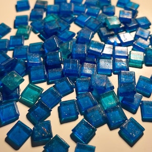 May include: A collection of small, square, blue glass tiles with a slightly iridescent finish. The tiles are scattered on a white surface.