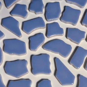 Könnte beinhalten: Eine Ansammlung unregelmäßig geformter, hellblauer Keramikfliesen. Jede Fliese hat eine glänzende Oberfläche und unterschiedliche Größen, mit einem leicht cremefarbenen Rand. Die Fliesen sind vor einem weißen Hintergrund verstreut.