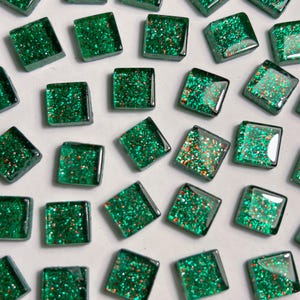 Peut inclure: Collection de carreaux de verre vert émeraude carrés avec des paillettes incrustées. Les carreaux sont dispersés sur une surface blanche, chacun reflétant la lumière. Les paillettes ajoutent un effet scintillant.