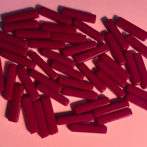 May include: A scattering of small, rectangular, magenta-coloured beads on a pale pink surface. The beads are smooth and evenly coloured, with a slight sheen. The arrangement is random, suggesting a craft supply or decorative element.