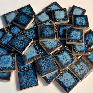 May include: A collection of square ceramic tiles with a dark blue glaze and a lighter blue, floral-like pattern in the center. The tiles have a slightly raised edge and a glossy finish, creating a textured appearance. The tiles are scattered across a white surface.
