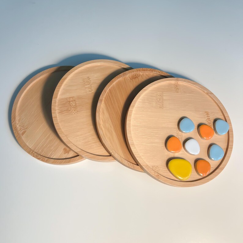 Op de afbeelding: Vier ronde houten schalen met een lichtbruine afwerking. De bovenste schaal heeft negen kleurrijke keramische stenen in tinten blauw, oranje, geel en wit.