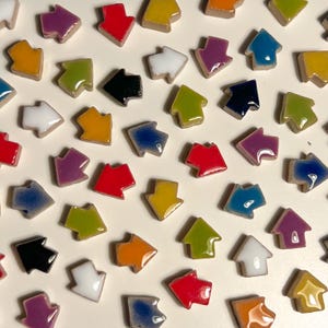 May include: Assortment of colorful ceramic arrow tiles in various colors, including red, blue, green, yellow, purple, and white. The tiles are arranged in a random pattern on a white background.