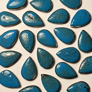 May include: A collection of turquoise teardrop-shaped ceramic beads with a speckled texture and a metallic silver border. The beads are arranged on a white surface, showcasing their vibrant color and unique design. These beads are ideal for jewelry making.