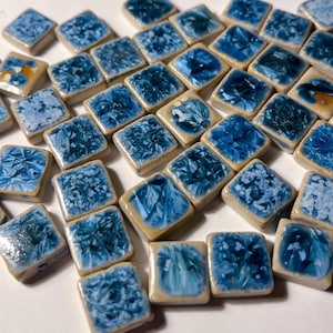 May include: A collection of square ceramic tiles with a light beige border and a glossy, blue crystalline pattern. The tiles vary in shades of blue, creating a textured and visually appealing mosaic effect. The tiles are arranged closely together.