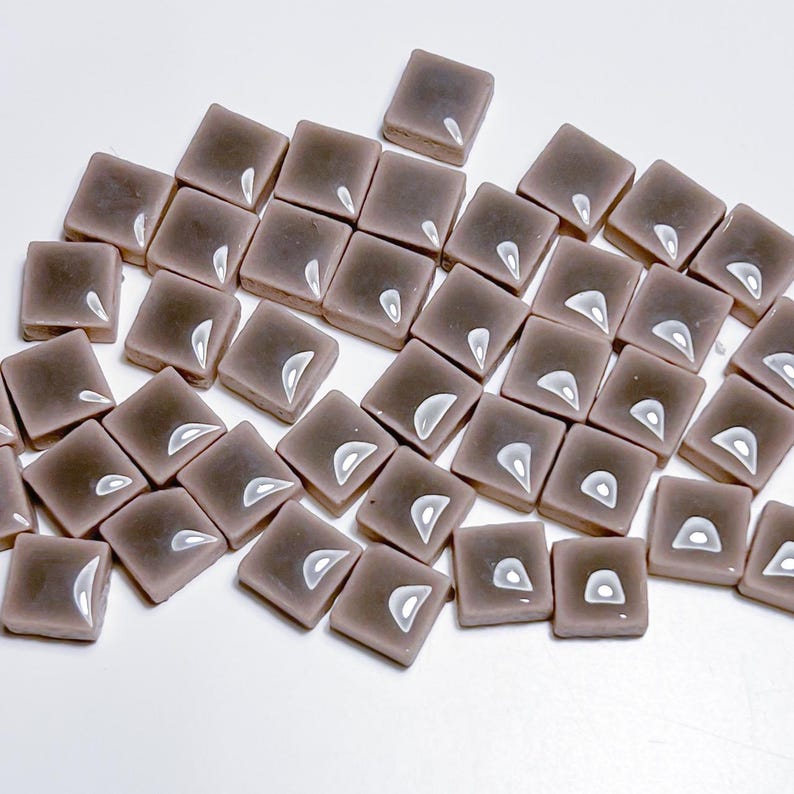 May include: A collection of small, square, mauve-coloured tiles with a glossy finish. Each tile has a white, teardrop-shaped design. The tiles are scattered on a white surface, suggesting they are for a mosaic or craft project.