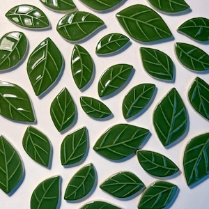 May include: A collection of glossy, green ceramic leaf tiles. Each leaf has a detailed vein pattern and a smooth, reflective surface. The leaves vary in size and are arranged on a white background, creating a natural, decorative element.