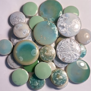 Puede incluir: Una colección de azulejos de cerámica circulares en varios tonos de verde, azul verdoso y blanco. Los azulejos presentan acabados lisos y brillantes, con algunos mostrando patrones cristalinos intrincados, tonos pastel sólidos y degradados.