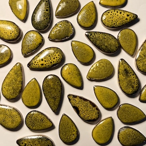 Puede incluir: Un surtido de piezas de cerámica en forma de lágrima en tonos amarillo y verde oliva. Las piezas tienen un patrón moteado y un acabado brillante, creando una textura única. La disposición sugiere una colección de elementos decorativos o suministros para manualidades.