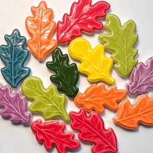 Op de afbeelding: Een verzameling kleurrijke keramische eikenbladdecoraties in verschillende tinten, waaronder rood, oranje, geel, groen, blauw en paars. Elk blad heeft gedetailleerde nerven en een glanzende afwerking. Deze decoratieve bladeren zijn ideaal voor herfstthema's.