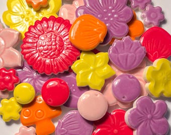 Op de afbeelding: Een verzameling kleurrijke keramische kralen en versieringen in verschillende bloem- en organische vormen. De collectie bevat rode, gele, oranje, roze en paarse bloemen, samen met ronde en schelpvormige kralen. Deze decoratieve elementen zijn ideaal voor knutselen en sieraden maken.