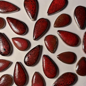 May include: An overhead shot displays numerous teardrop-shaped ceramic pieces. The pieces vary in shades of red and brown, with some featuring a speckled pattern. The glossy finish reflects light, creating highlights. The objects are arranged on a white surface.