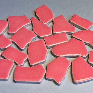 May include: A collection of irregularly shaped, pink ceramic tiles with a glossy finish and gray edges. The tiles are scattered across a light gray surface, showcasing their varied forms and textures. These tiles are suitable for mosaic art projects.