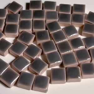 Op de afbeelding: Een verzameling kleine, vierkante, grijze keramische tegels. De tegels hebben een glanzende afwerking en zijn verspreid over een wit oppervlak.