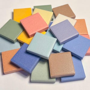 以下が含まれることがあります： 黄色、ピンク、緑、青など、さまざまなパステルカラーの小さな正方形のタイルが集まっています。 タイルは山のように配置され、一部は重なっています。 タイルは固い素材でできているようです。