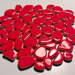 Könnte beinhalten: Eine Sammlung von glänzenden, unregelmäßig geformten Keramikfliesen in leuchtendem Rot. Die Fliesen sind in verschiedenen Größen und Formen und ähneln Kieselsteinen.