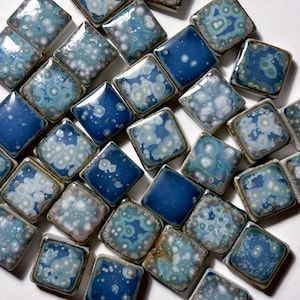 May include: An assortment of square ceramic tiles in shades of blue and teal. Some tiles have a solid blue glaze, while others feature a speckled pattern. The tiles are arranged closely together, creating a textured surface.
