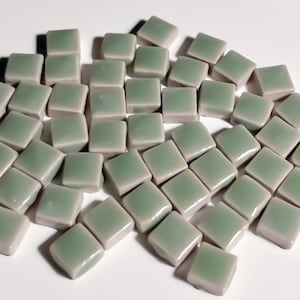 May include: A collection of small, square, sage green tiles. The tiles have a glossy finish and are arranged in a scattered pattern. These tiles are suitable for mosaic projects or decorative applications.