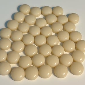 Puede incluir: Primer plano de numerosas cuentas redondas de color crema. Las cuentas están muy juntas, llenando la imagen. Cada cuenta tiene una superficie lisa, ligeramente abovedada, que refleja la luz. La disposición crea un patrón uniforme y táctil.