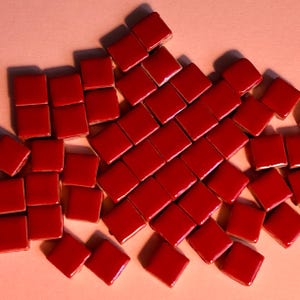 Peut inclure: Une collection de petites tuiles carrées en céramique rouge éparpillées sur une surface rose pâle. Les tuiles sont disposées en un motif lâche et organique.