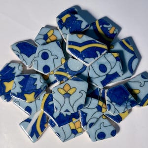 May include: A collection of broken ceramic tiles with a light blue background and intricate designs in dark blue and yellow. The tiles are irregularly shaped, with floral and geometric patterns. Ideal for mosaic art or craft projects.