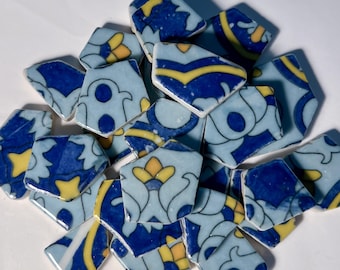 Op de afbeelding: Een verzameling gebroken keramische tegels met een lichtblauwe achtergrond en ingewikkelde ontwerpen in donkerblauw en geel. De tegels zijn onregelmatig van vorm, met bloemen- en geometrische patronen. Ideaal voor mozaïekkunst of knutselprojecten.