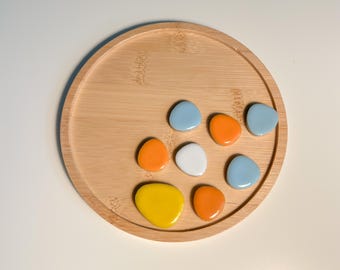 May include: A wooden tray with seven ceramic stones in various colors, including blue, orange, yellow, and white. The stones are arranged in a circular pattern on the tray.