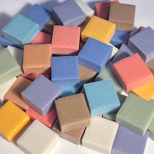 May include: A collection of small, square mosaic tiles in various pastel colors, including blue, pink, yellow, and green. The tiles are arranged in a pile, showcasing their smooth, matte surfaces. These tiles are suitable for craft projects.